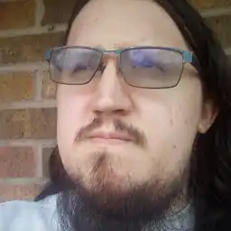 White man in his 20s, wearing shaded glasses and sporting a thick scraggly beard.